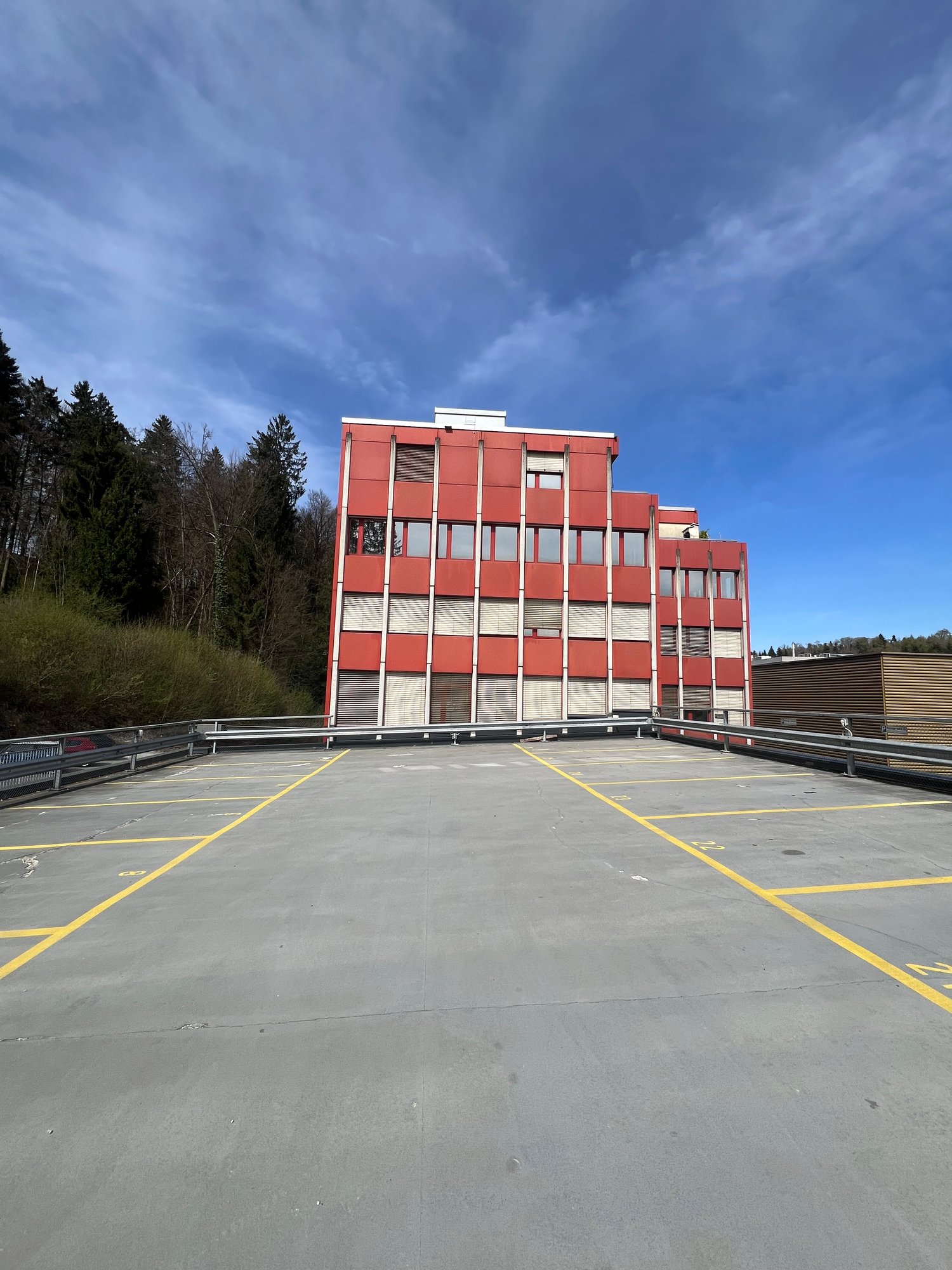 A multi-story red and white building with a large parking lot in the foreground. The building appears to be a commercial or industrial structure surrounded by trees and a cloudy blue sky.