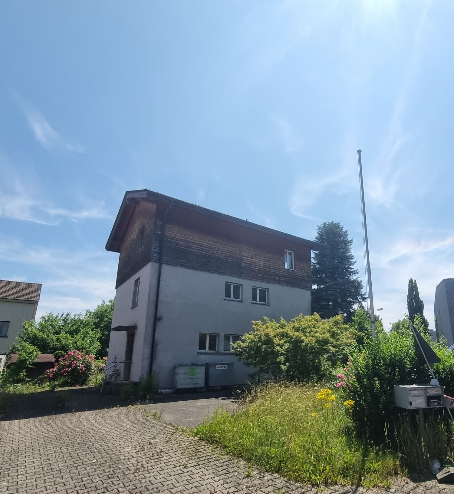 2-story house with wooden siding on the upper floor, gray exterior walls, large windows, surrounded by trees and vegetation, paved driveway or walkway leading to the house
