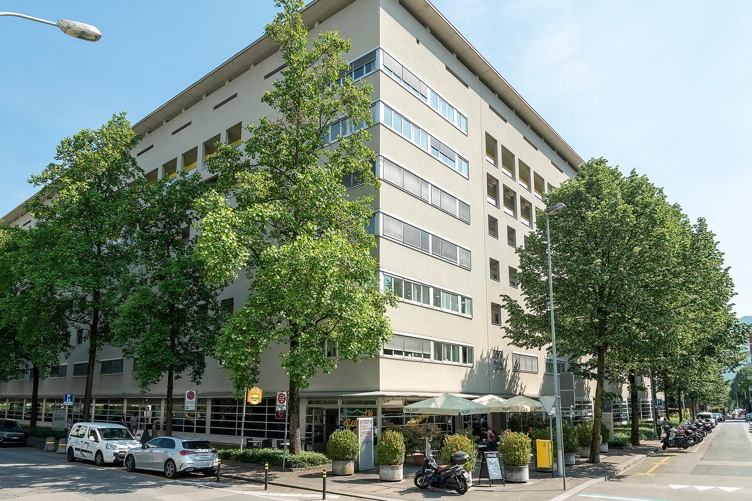 An apartment building with many windows, a road, some cars parked in front, trees, a street light, a trash can, a few people sitting under umbrellas, and a motorcycle parked on the side.