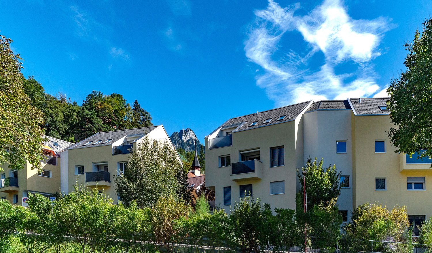 Two multi-story buildings with balconies, yellow exterior, trees, mountains in background, road