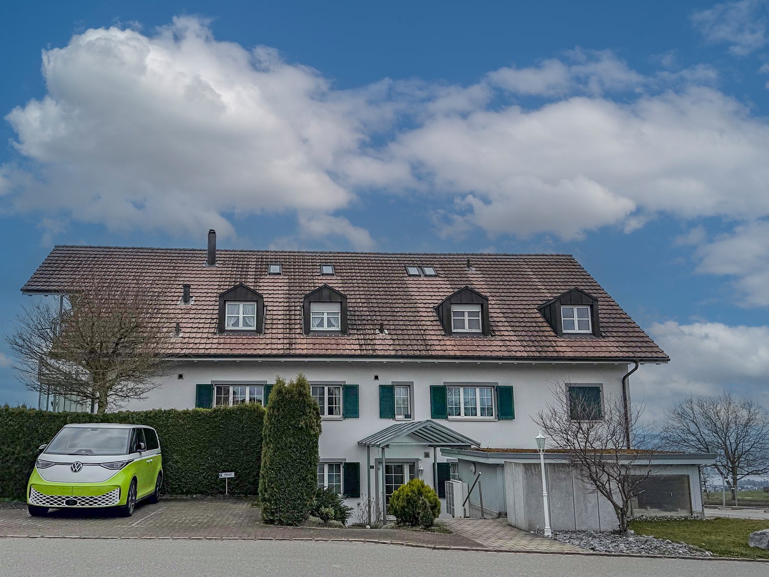 2 story house with a tiled roof, white exterior walls, green shutters, and a garage. There is a green Volkswagen ID.Buzz electric vehicle parked in front of the house.