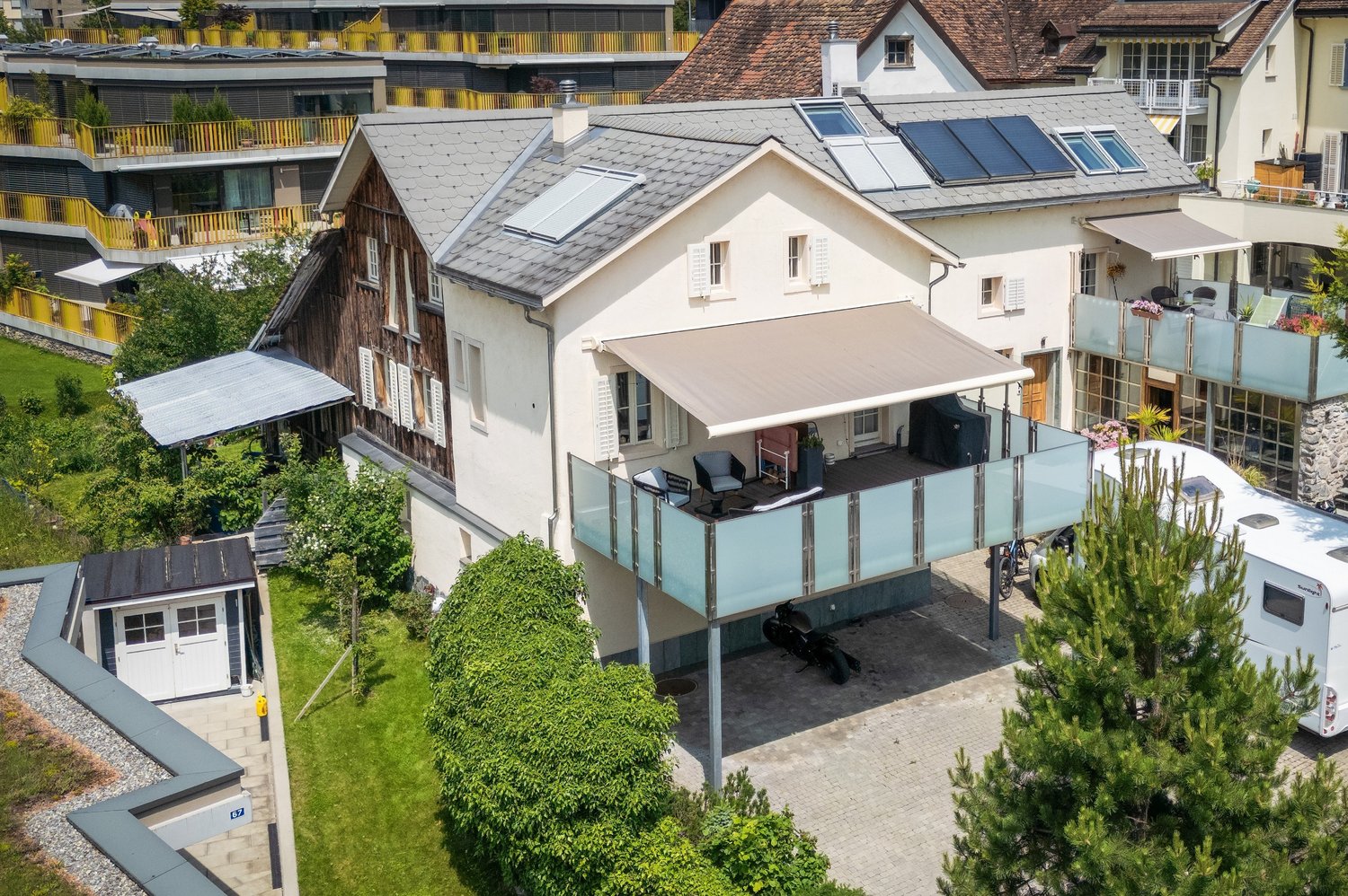A white house with solar panels, a large balcony with awning, and a covered parking area with a motorcycle and a white trailer