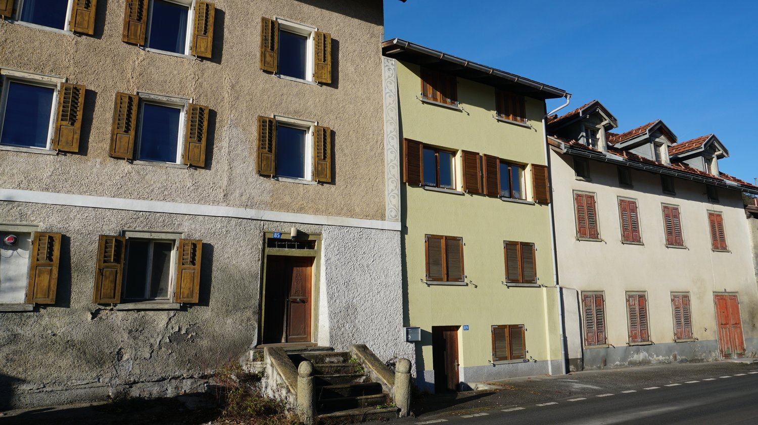 Three buildings side by side, each with multiple windows, wooden shutters, and entrances, made of concrete.