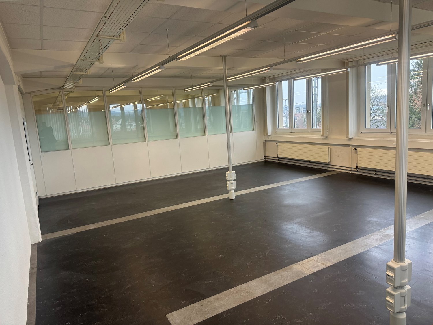 Empty room with white walls, black floor, glass partitions, windows, and overhead lighting.