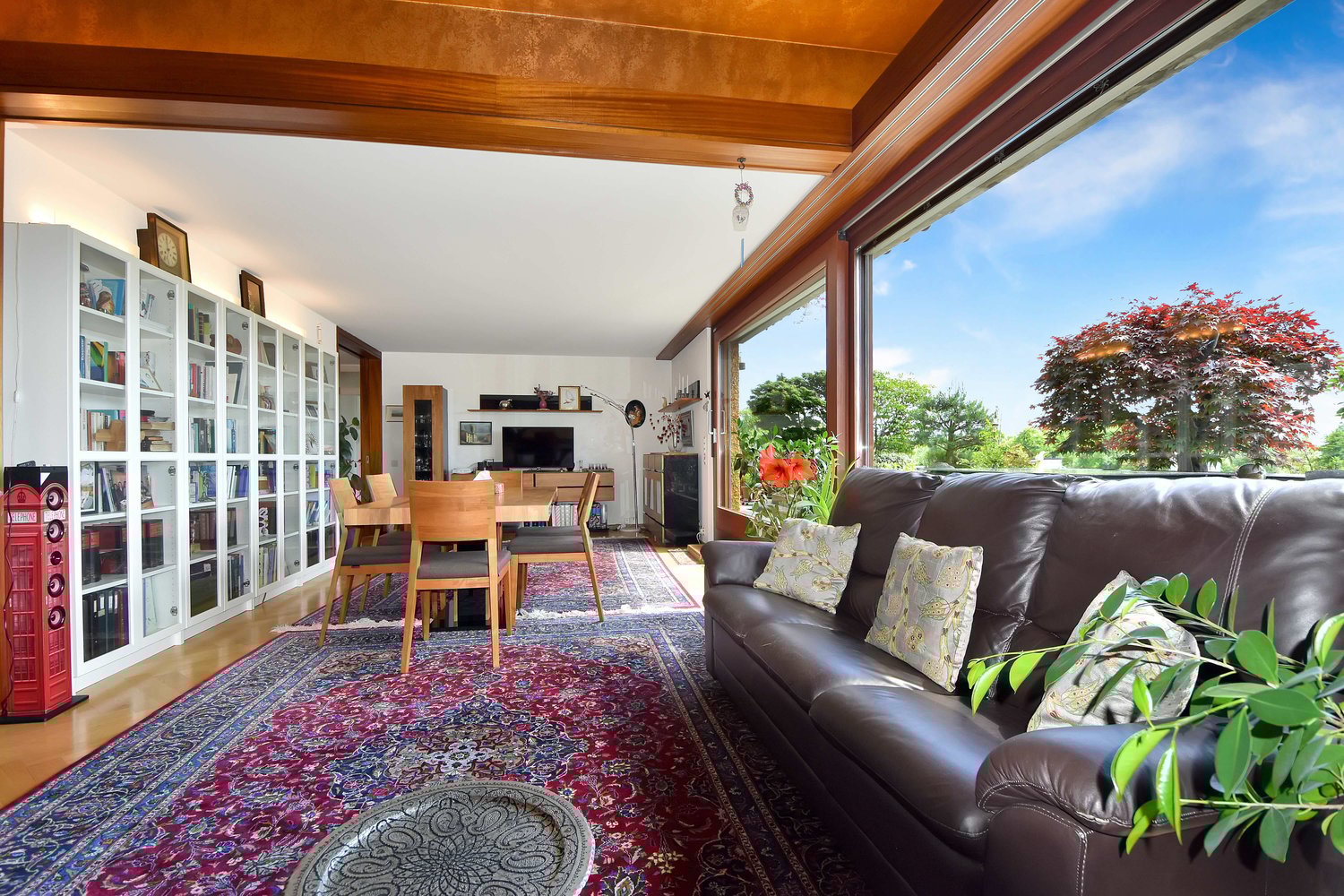 Spacious living room with large windows overlooking a scenic outdoor view, wooden ceiling beams, built-in bookshelves, leather sofa, dining table and chairs, colorful patterned rug