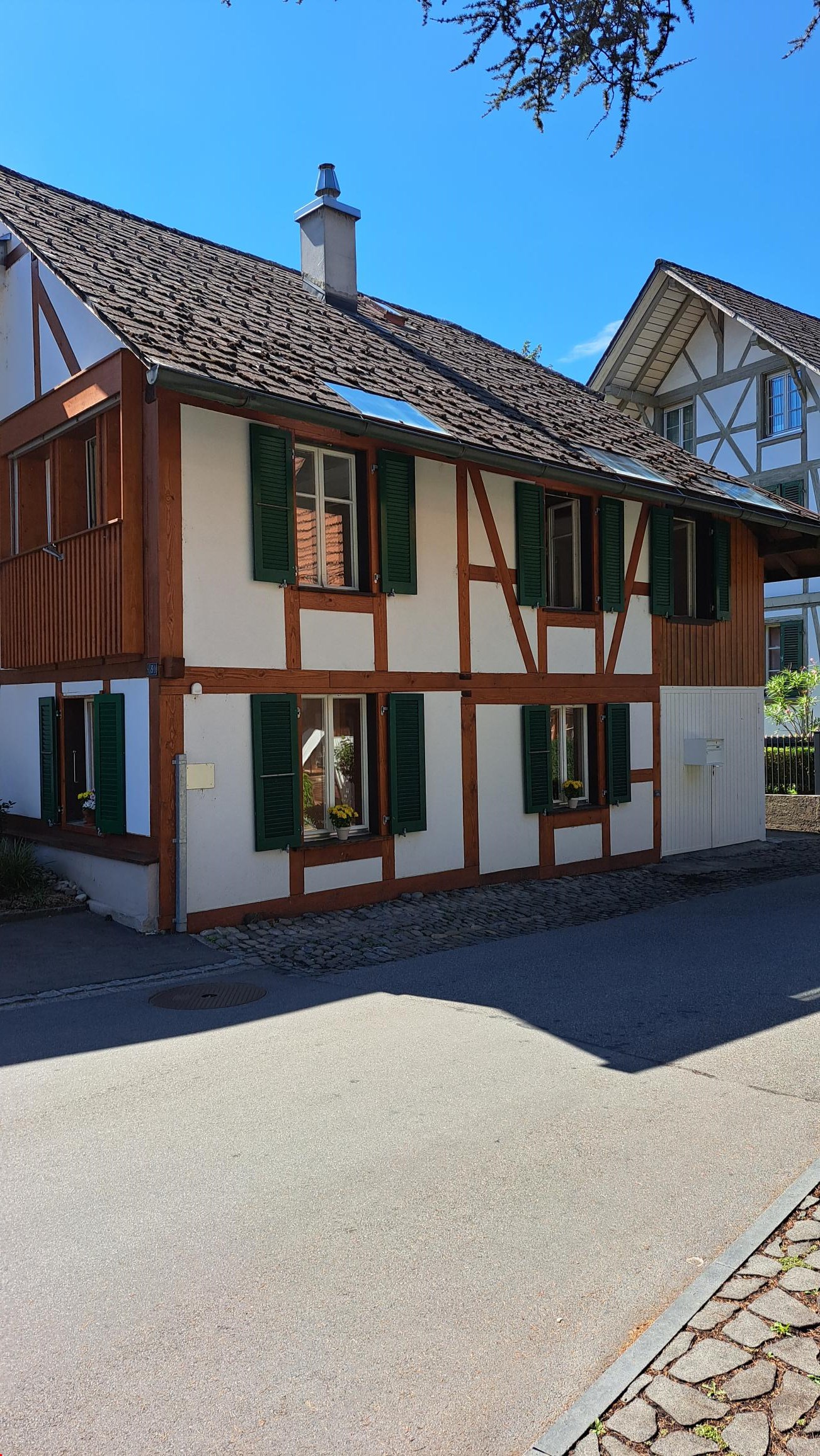 Raised ground floor, brown roof, wooden beams, windows with green shutters, solar panels on the roof, small garden at the front.