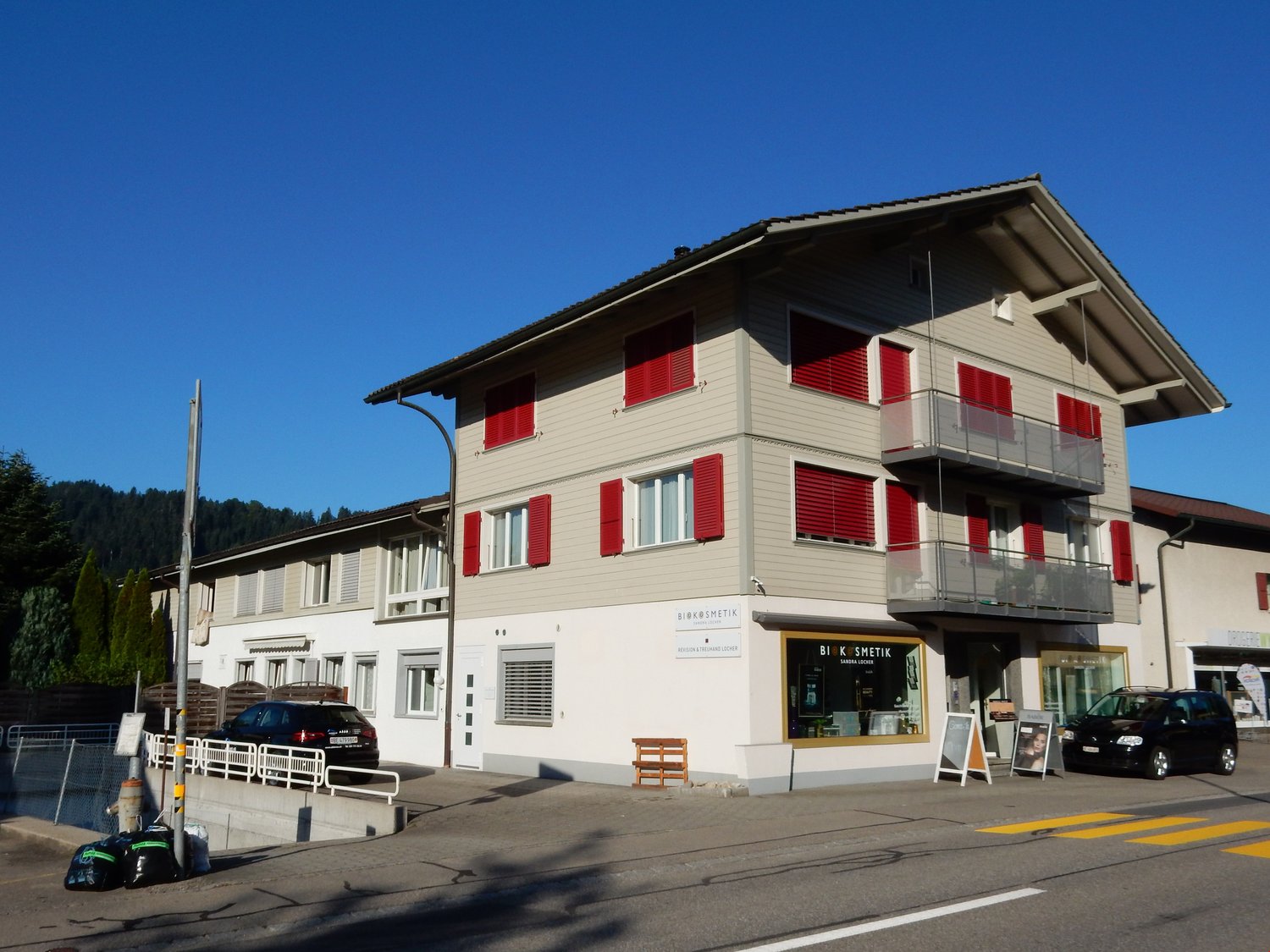 Apartment building, red shutters, gray and white walls, first floor shop, two cars parked in front, trees in the background, sign with text "BIK ESMETI"