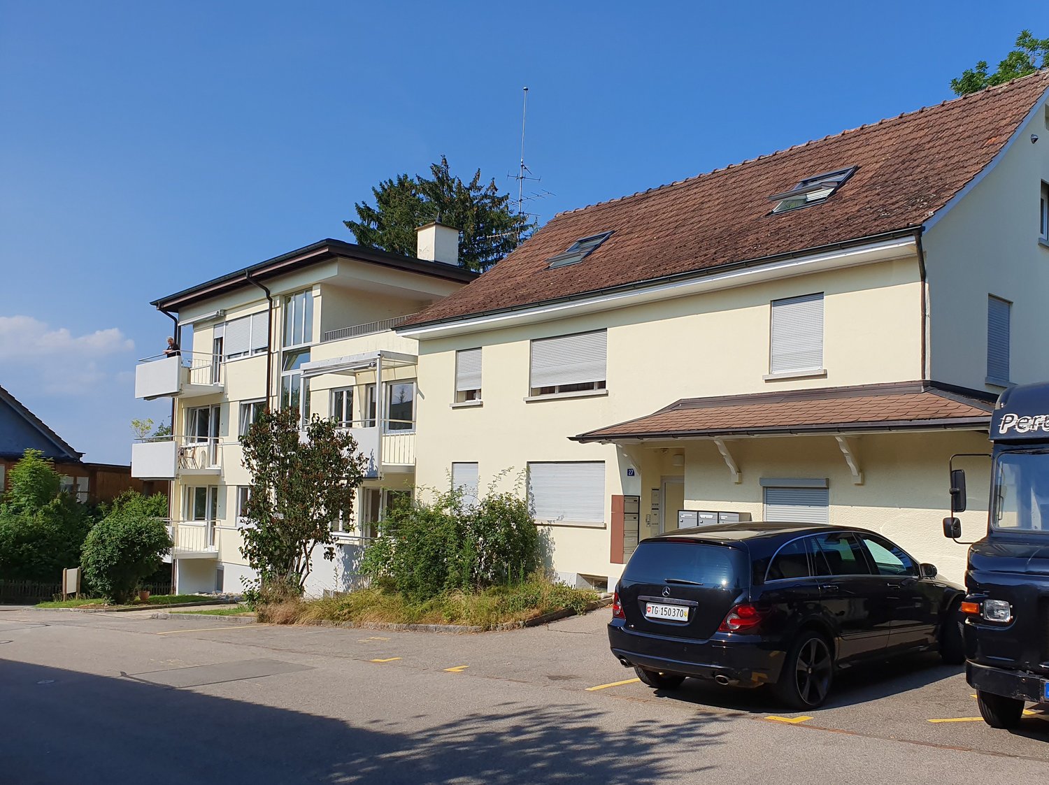 Building with white walls and a brown roof, balconies on the second floor, parking area with a car parked.