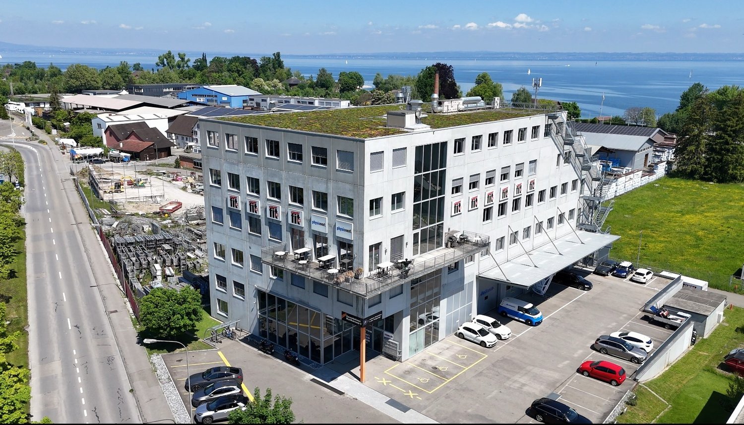 Multi-story commercial building with a green roof, located near a body of water with a view. The building has a parking lot in front and appears to be used for office or business purposes.