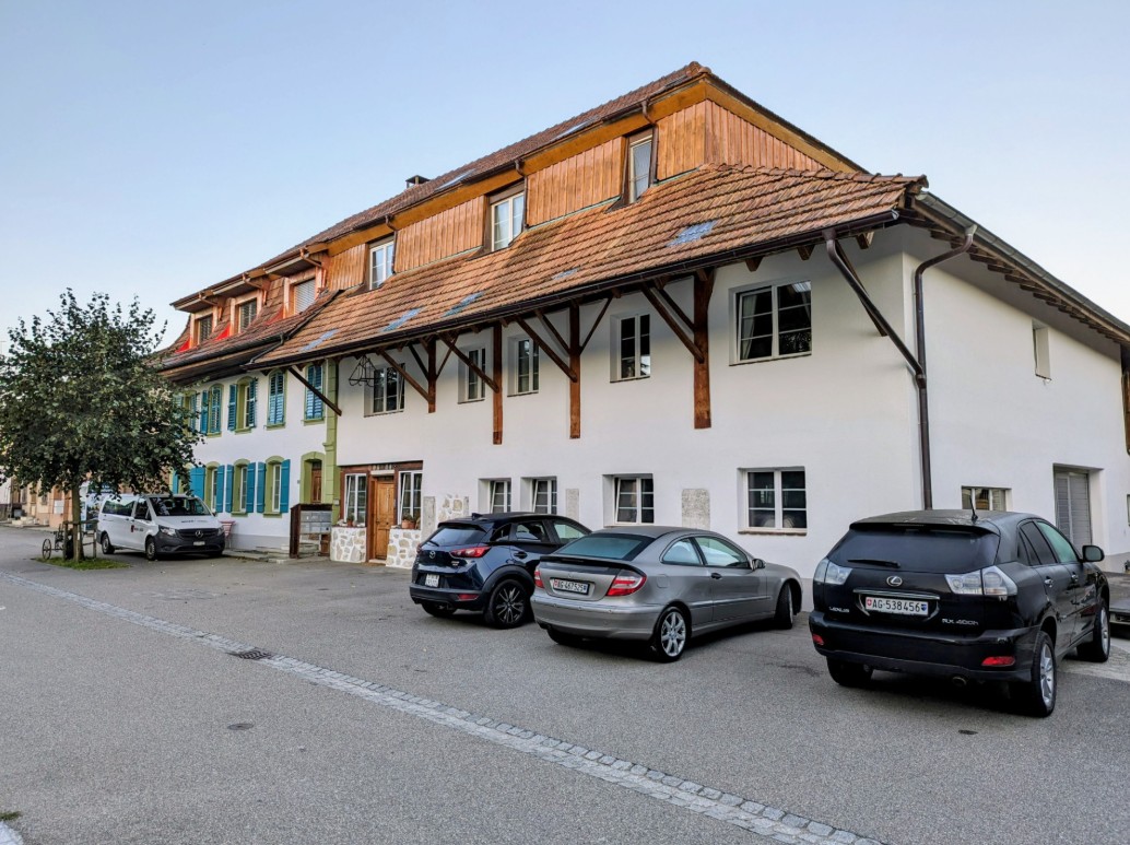 Large white building, red tiled roof, multiple windows, cars parked in front, brick base, wooden beams