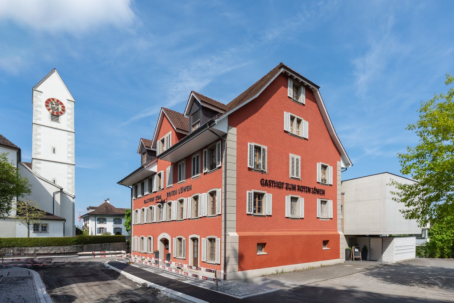 Red painted building, white trims, white windows with shutters, clock tower visible, two trees in front, beige curb