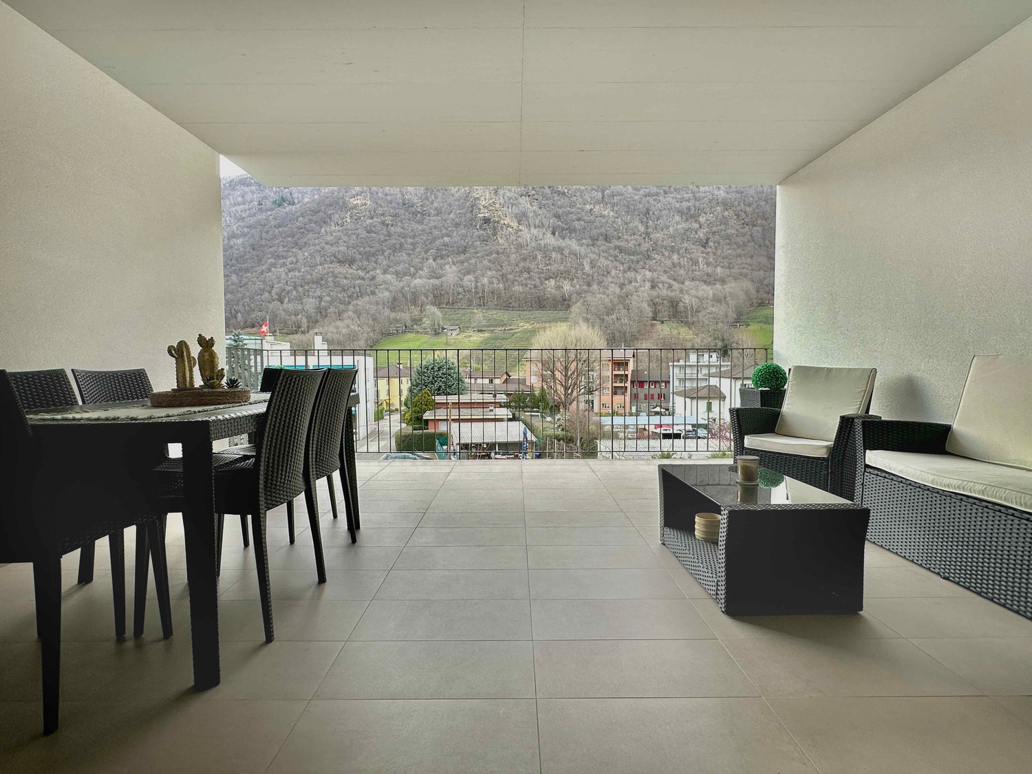 Spacious living room with large windows overlooking a scenic mountain landscape, black wicker furniture including a dining table and chairs, and a balcony or terrace area.