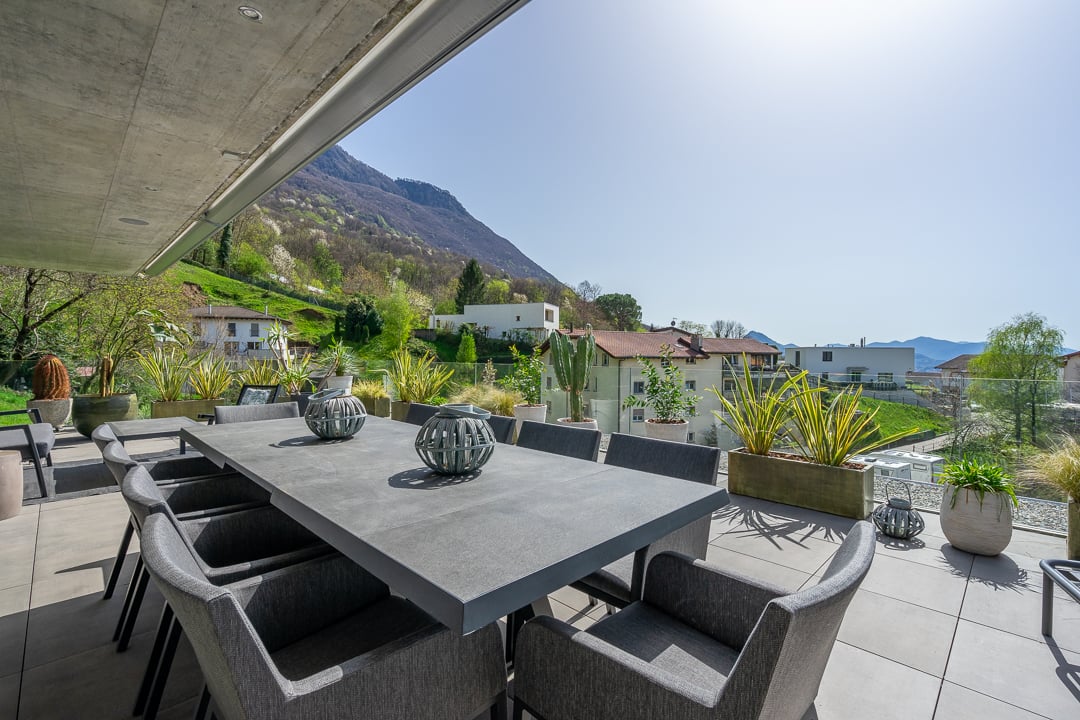 Spacious outdoor patio with a large concrete dining table and chairs, surrounded by potted plants and greenery. The patio overlooks a scenic mountain landscape in the background.