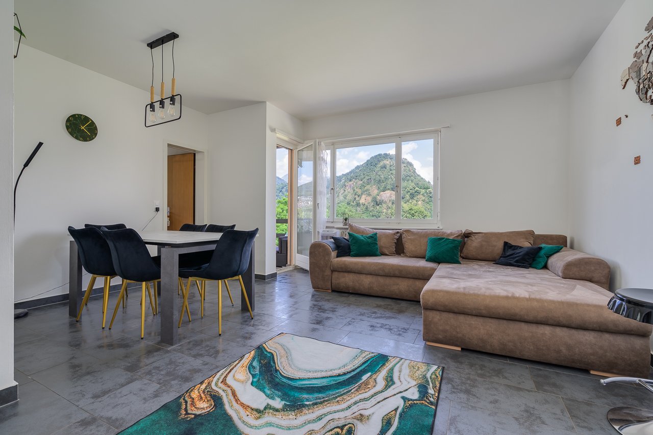 Spacious open-plan living area with large windows offering a scenic mountain view, a comfortable brown sofa, a dining table with black chairs, and a modern light fixture overhead.