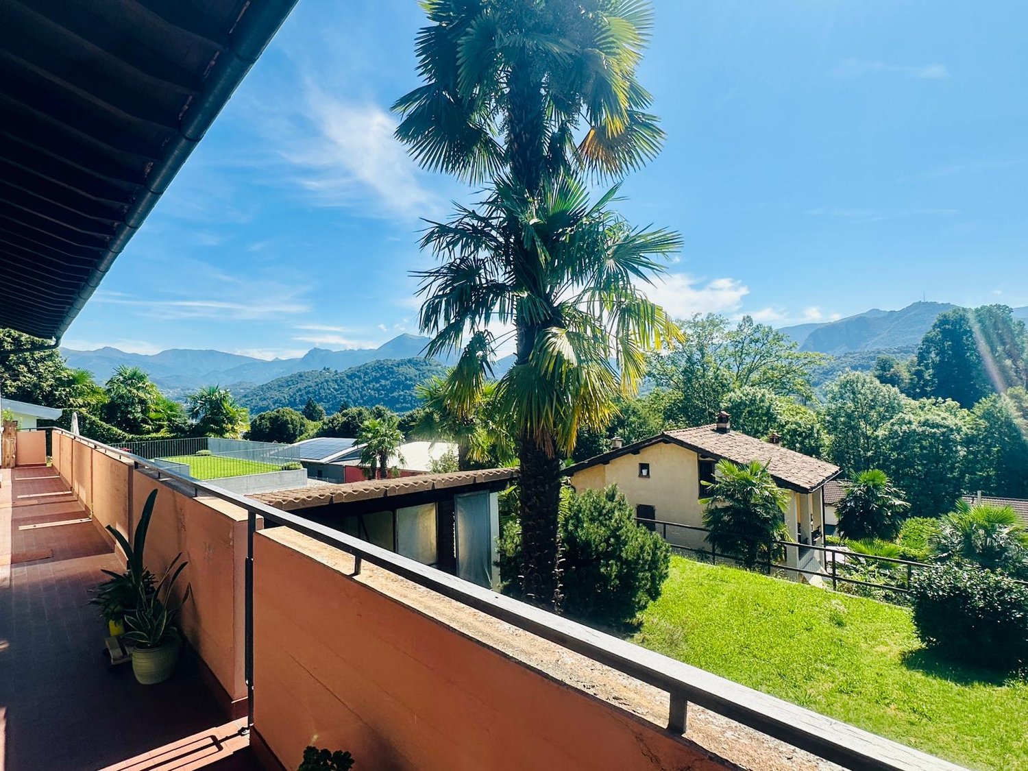 The image shows a residential property with a balcony or terrace overlooking a lush, green landscape. The property is surrounded by palm trees and other vegetation, with mountains visible in the background, indicating a scenic, tropical setting.