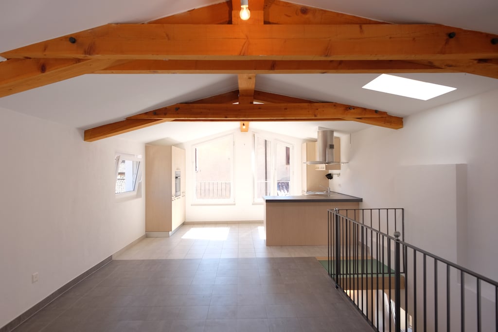 Empty room with white walls and tiled floor, wooden beams ceiling, windows, and kitchen.