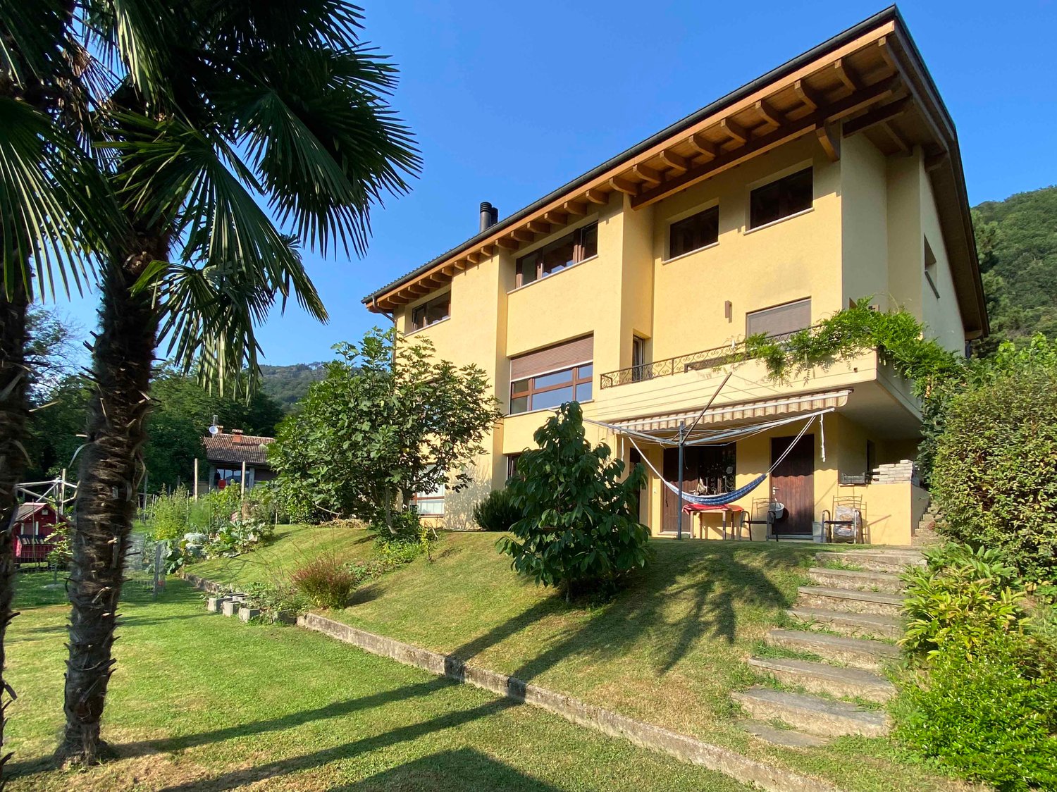 2-story yellow house with wooden roof, balconies, surrounded by lush greenery and palm trees, raised ground floor with steps leading to the entrance
