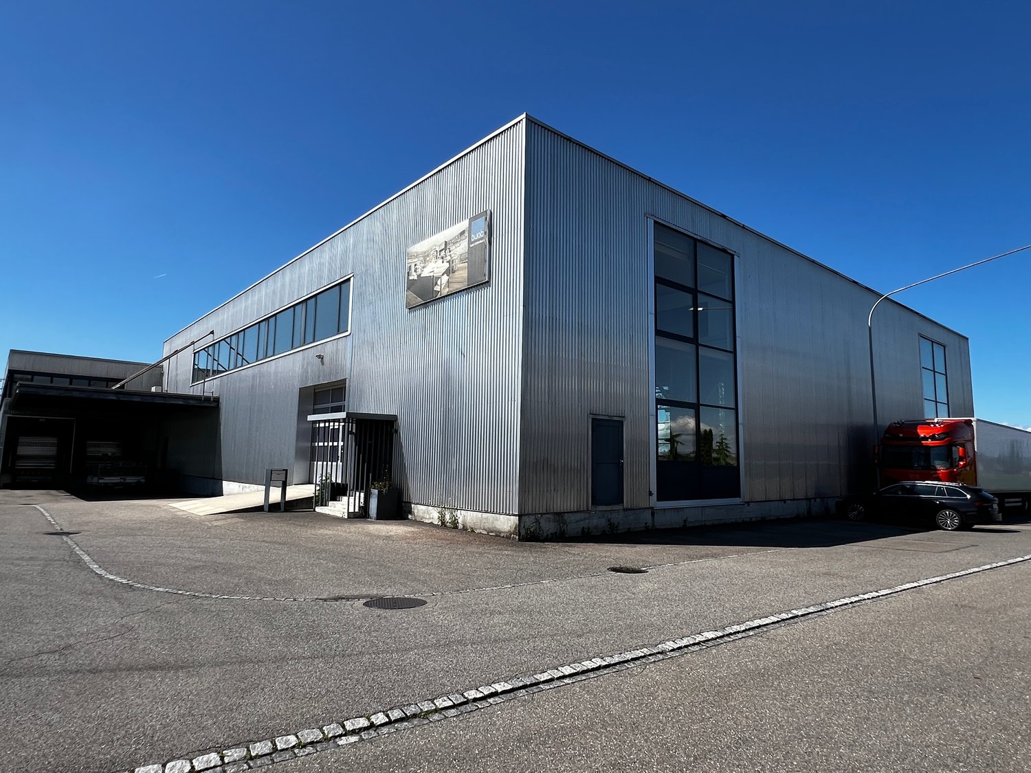 Industrial building with metal siding, large windows, a sign on the wall, a car parked in front, and a truck parked to the side.