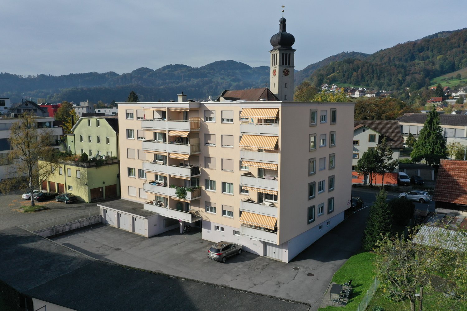 5 story building, multiple balconies, parking area, clock tower, nearby church, green hill in background