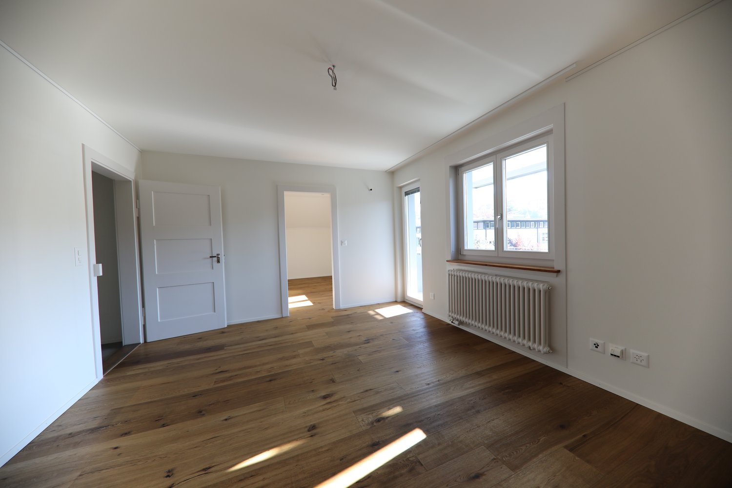 Spacious room with white walls, hardwood flooring, large windows, and a radiator. The room appears to be empty, ready for furnishing and decorating.