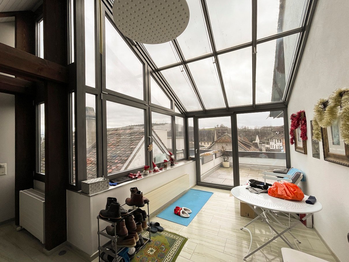 Room with large windows, a white table, a rug, and a shower head on the ceiling. The view outside the windows shows a snowy rooftop and cityscape.