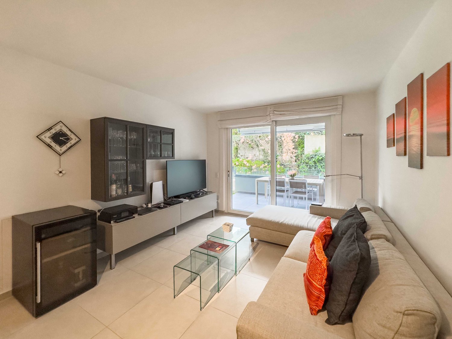 modern living room with beige couch, glass coffee tables, flat screen tv, wine glasses and bottles in the glass cabinet, balcony, white walls, tile floor