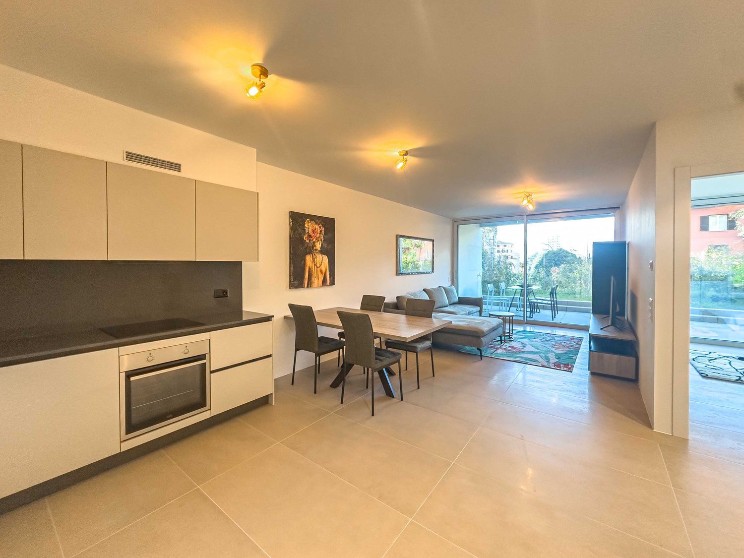 modern kitchen with black countertops, white cabinets, modern appliances, dining area with table and chairs, living area with couch, TV, large glass door leading to balcony, modern artwork