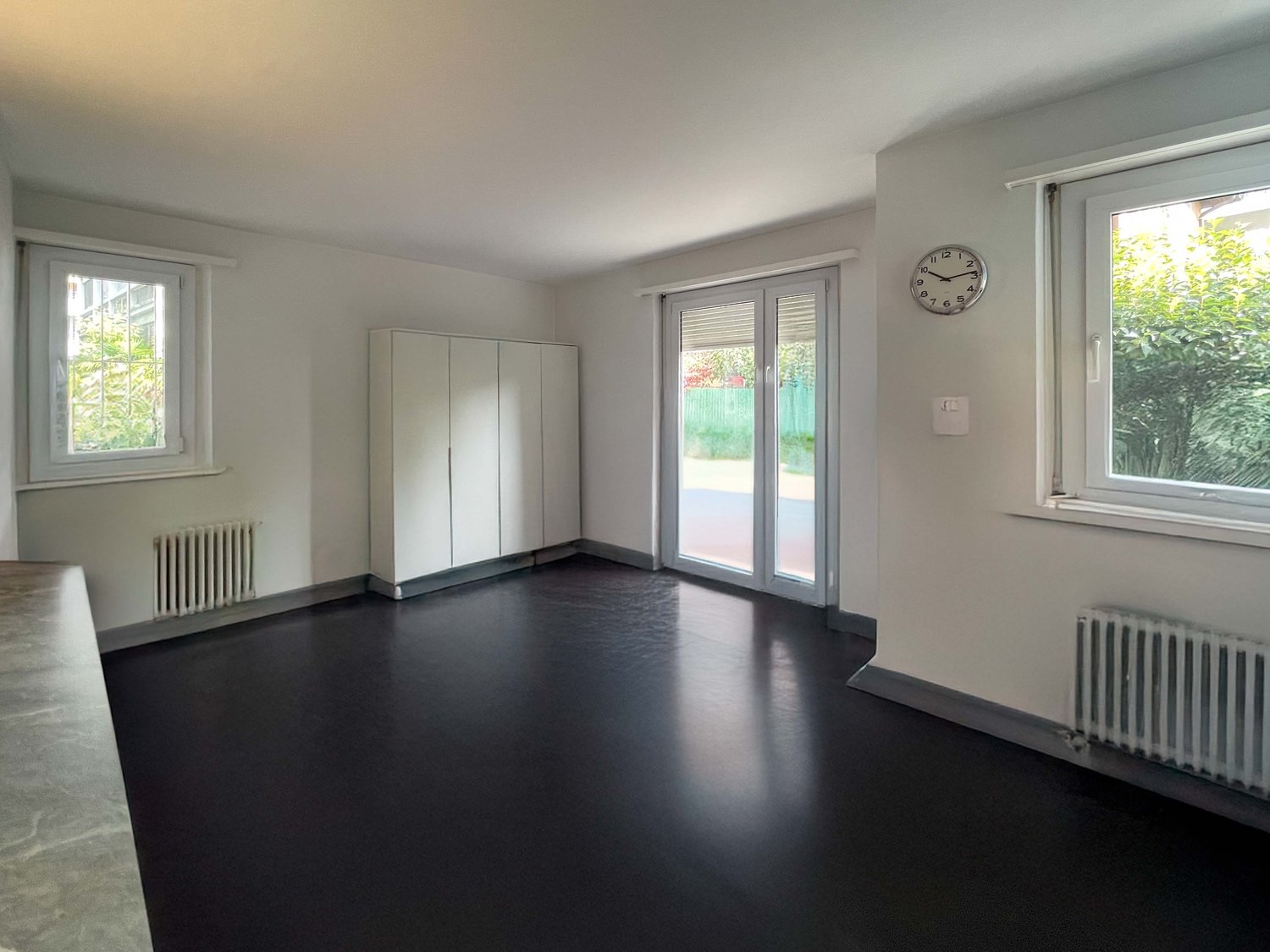 Empty room, white walls, black flooring, white sliding glass door, large windows, wall-mounted clock, two white radiators