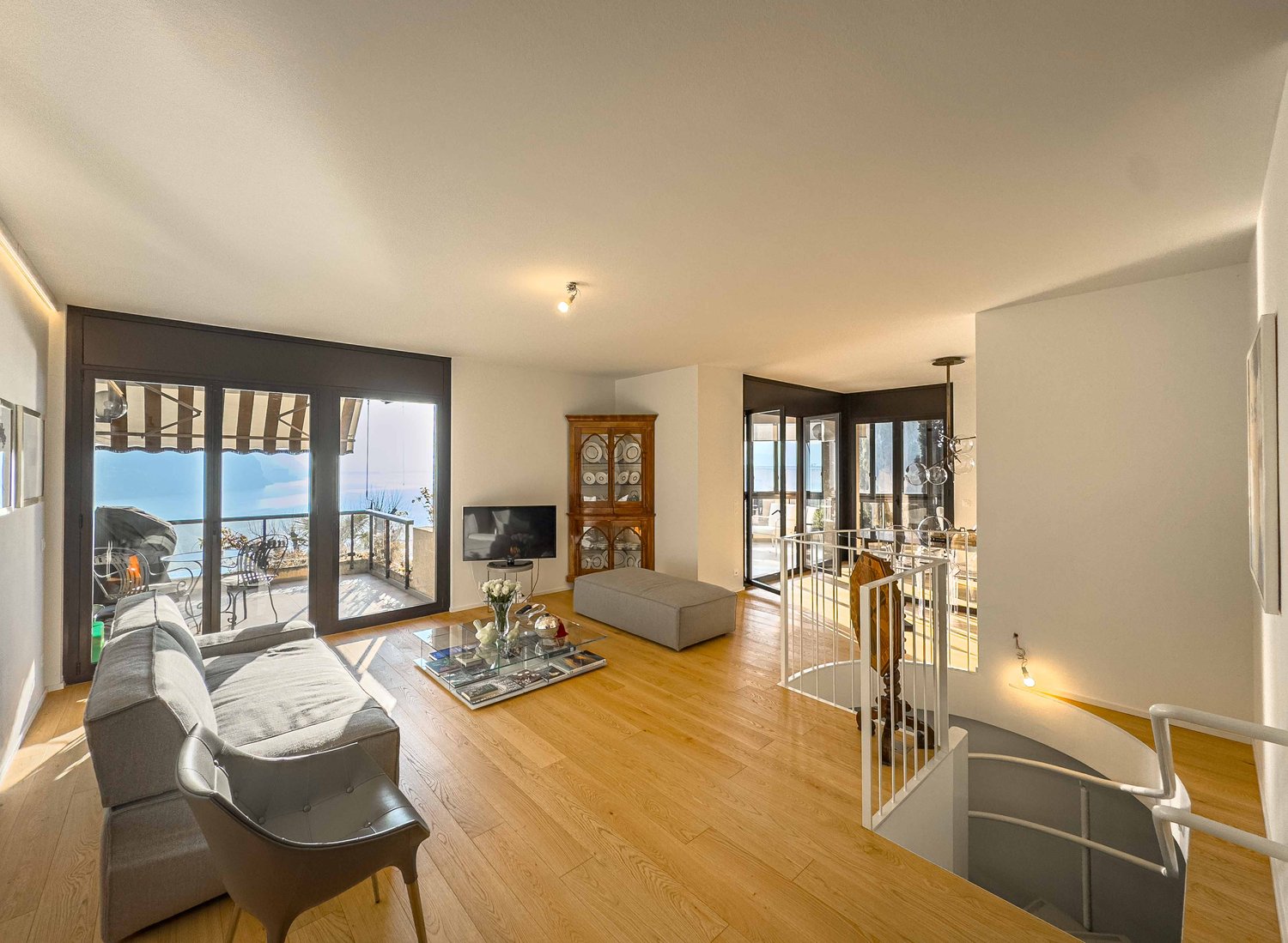 Modern living room with wooden floor, large sofa, glass coffee table, and glass balcony doors overlooking a view. TV mounted on wall, spiral staircase leading to upper level.