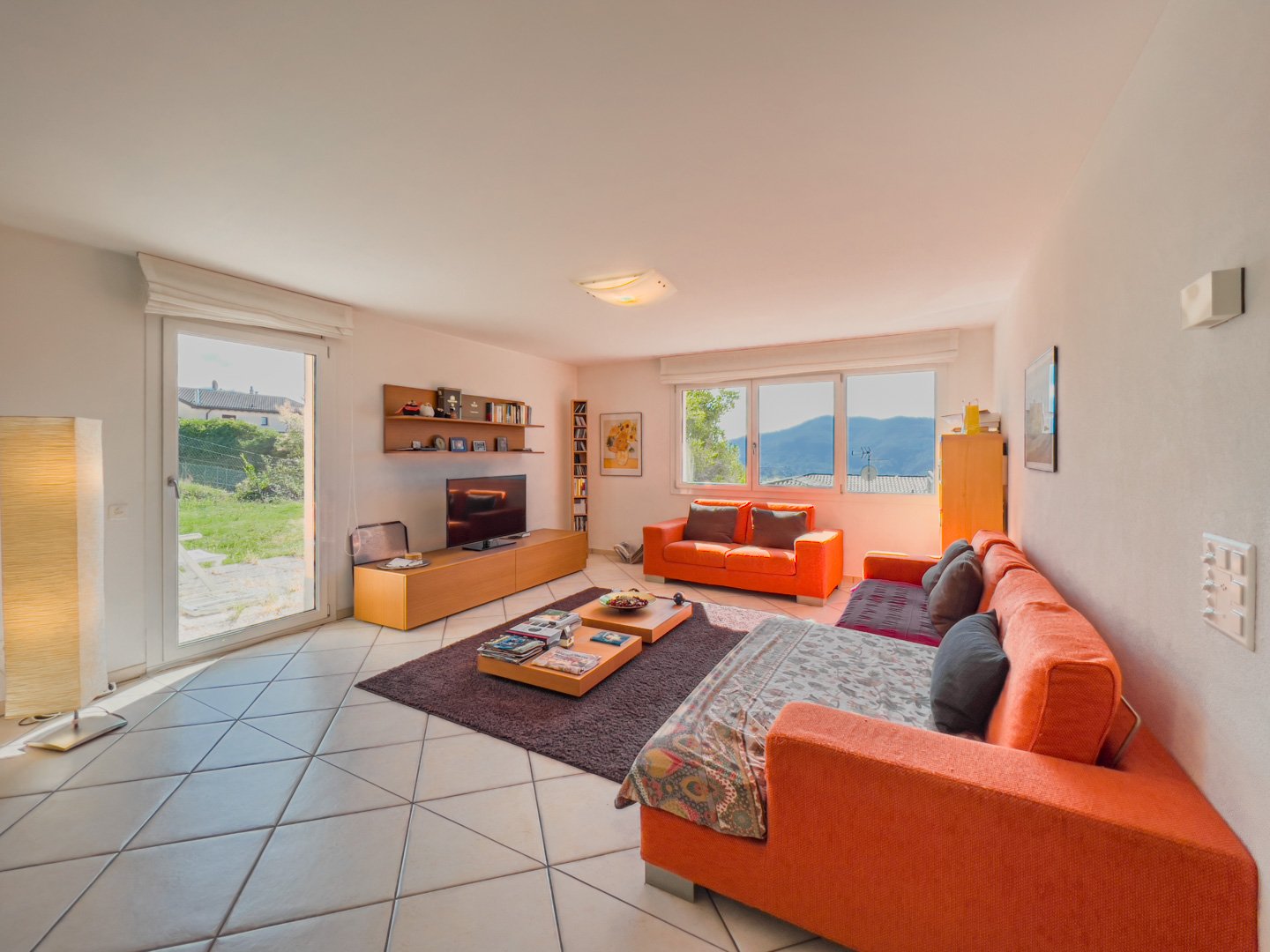 A modern living room with white walls and ceiling, large windows, tile floor, an orange sofa, center tables with books, a flat-screen TV on a TV cabinet, a wooden shelf with various items on it, and a view of the outdoors from the windows.
