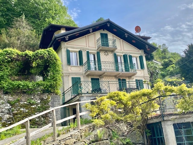 2-story house with a traditional Alpine-style architecture, featuring a sloped roof, wooden balconies, and green shutters. The house is surrounded by lush greenery and situated on a hillside overlooking a scenic landscape.