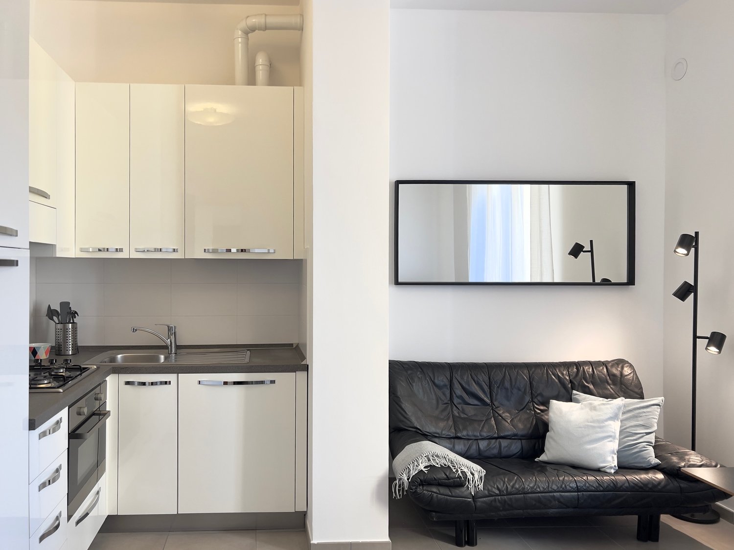 The image shows a small studio apartment with a kitchenette and a living area. The kitchenette has white cabinets, a sink, and a dishwasher. The living area has a black leather sofa, a large wall mirror, and a floor lamp.