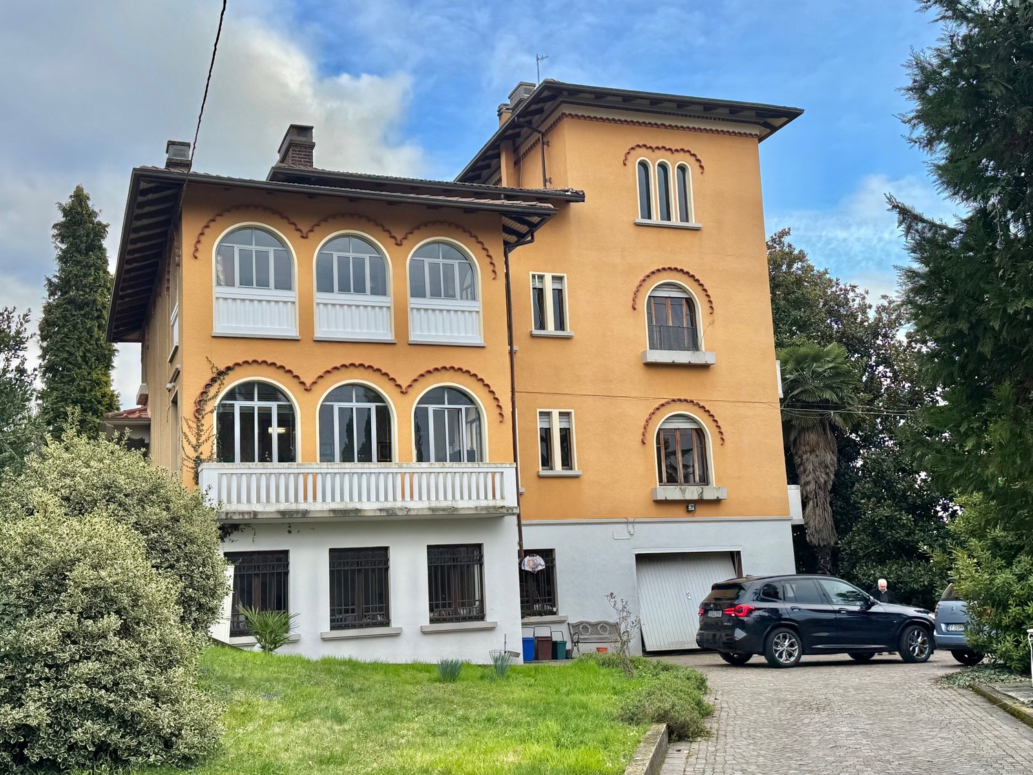 3-story yellow house with arched windows, decorative trim, and a balcony. The house has a garage and is surrounded by trees and greenery.