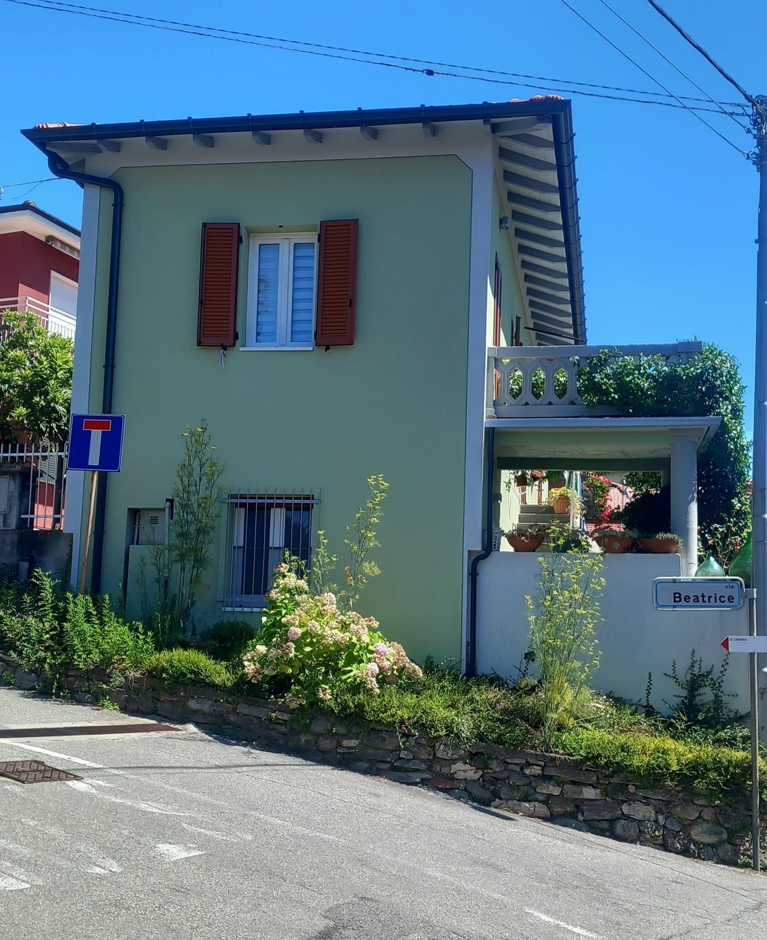 Two-story house with green exterior, red shutters, balcony, and landscaped garden with flowers and plants. The house has a street sign indicating the name 'Beatrice'.