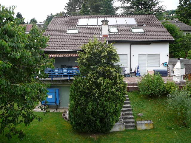 2-story house with a tiled roof, white exterior walls, and a balcony on the upper level. There is a garage attached to the side of the house. The house is surrounded by trees and greenery in the yard.