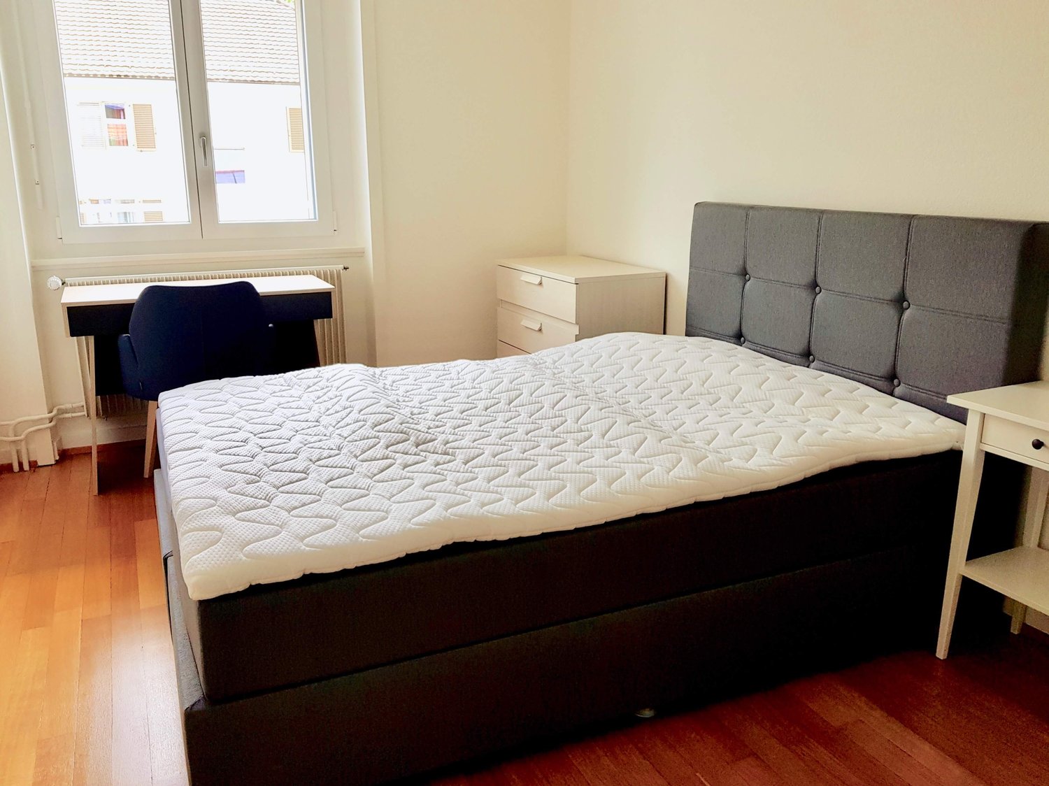 Empty bedroom with bed, desk, and chair. Wooden floor, white walls, window with curtains, wardrobe with drawers, and nightstands.
