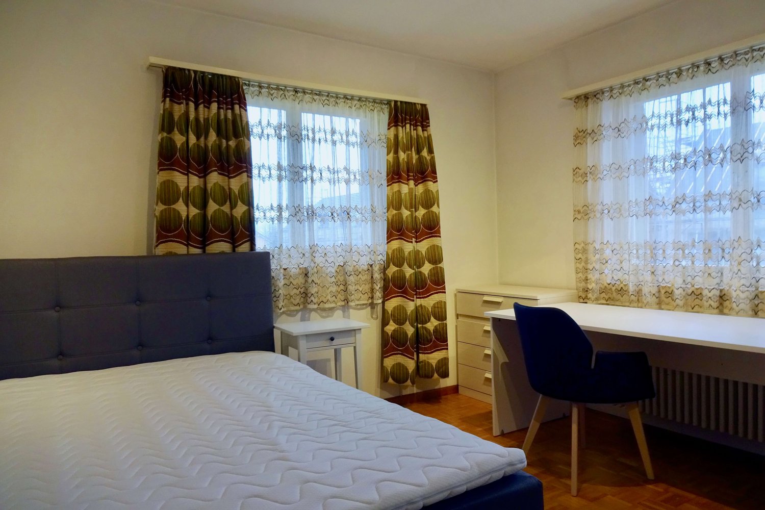 bed, white sheets, nightstand, desk, chair, wooden floor, curtains, radiator, cabinet