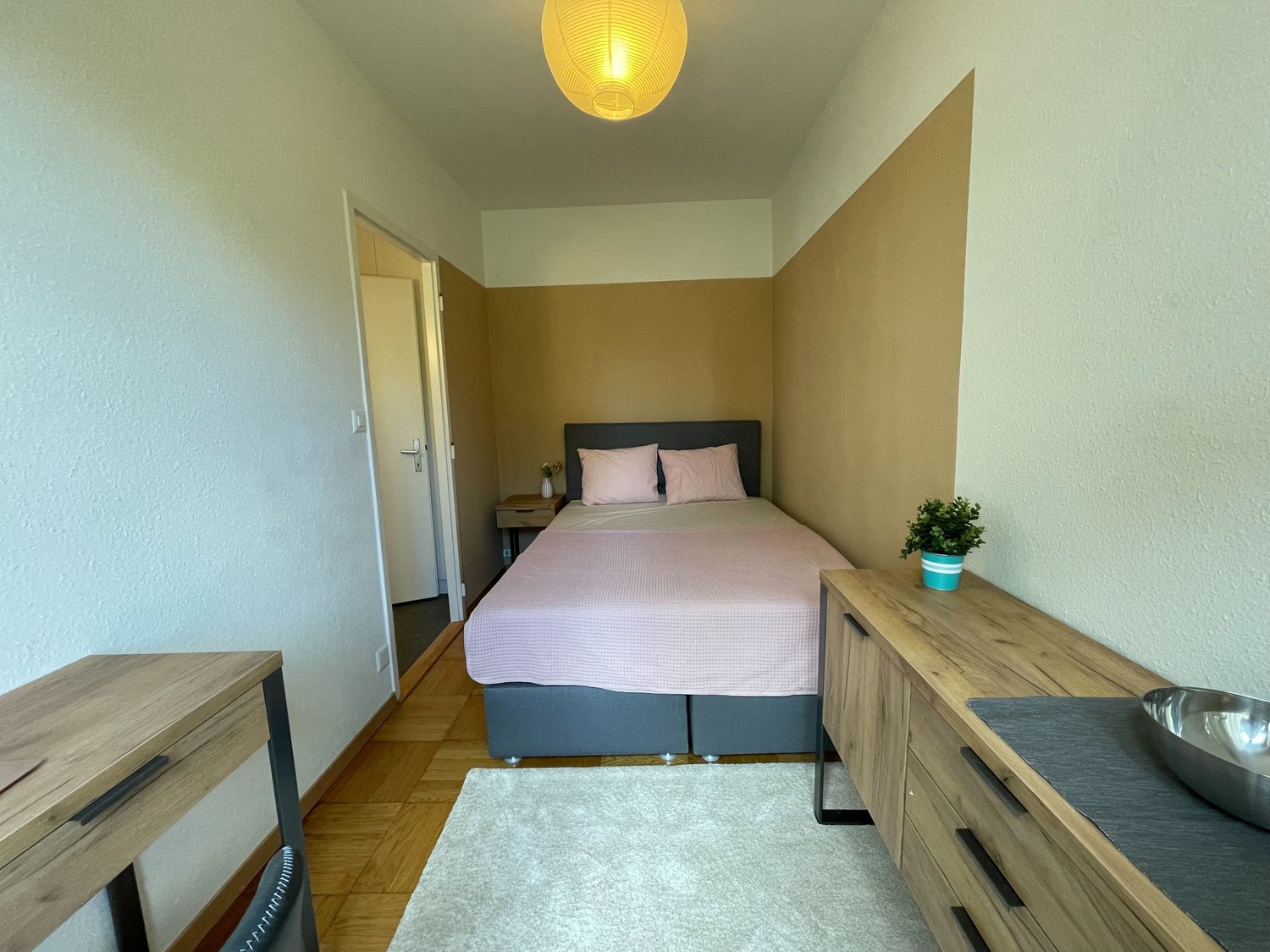 Bedroom with a bed, nightstand, lamp, desk, rug, plant, and wooden cabinet