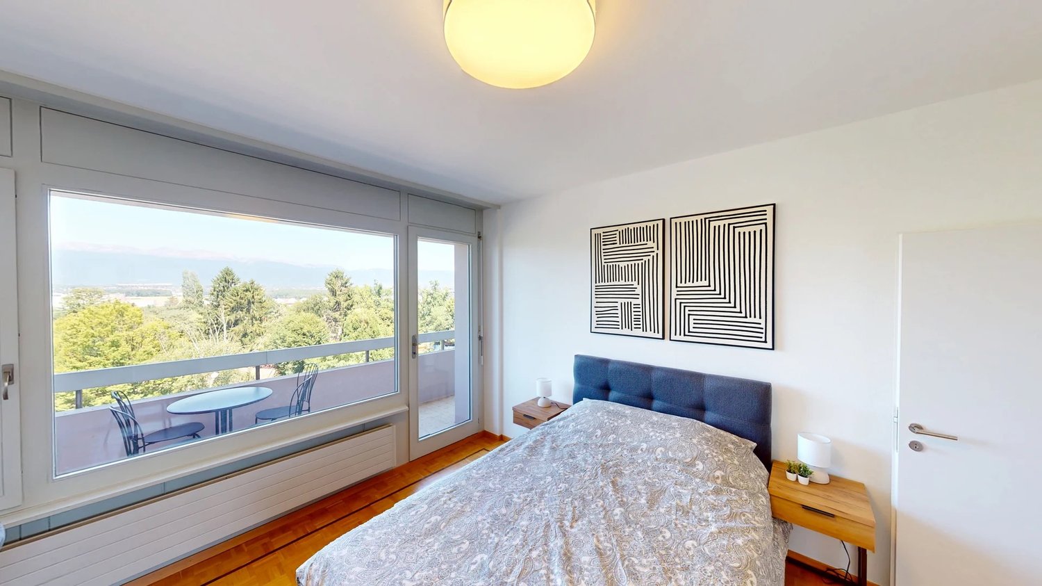 bed, nightstand, lamp, balcony with table and chairs, framed artwork on wall, large windows with view of trees