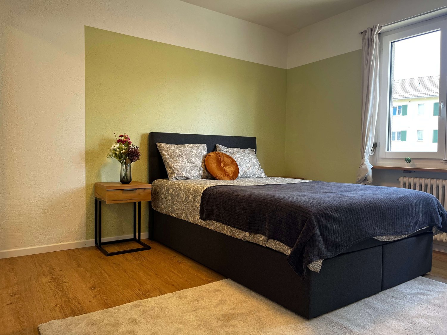 bed with pillows, flower vase, small wooden table, white rug, window with curtains, radiator