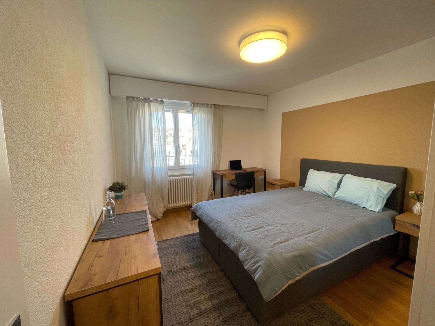 Bedroom with bed, wooden desk, lamp, window, radiator, rug, potted plant, cup and vase