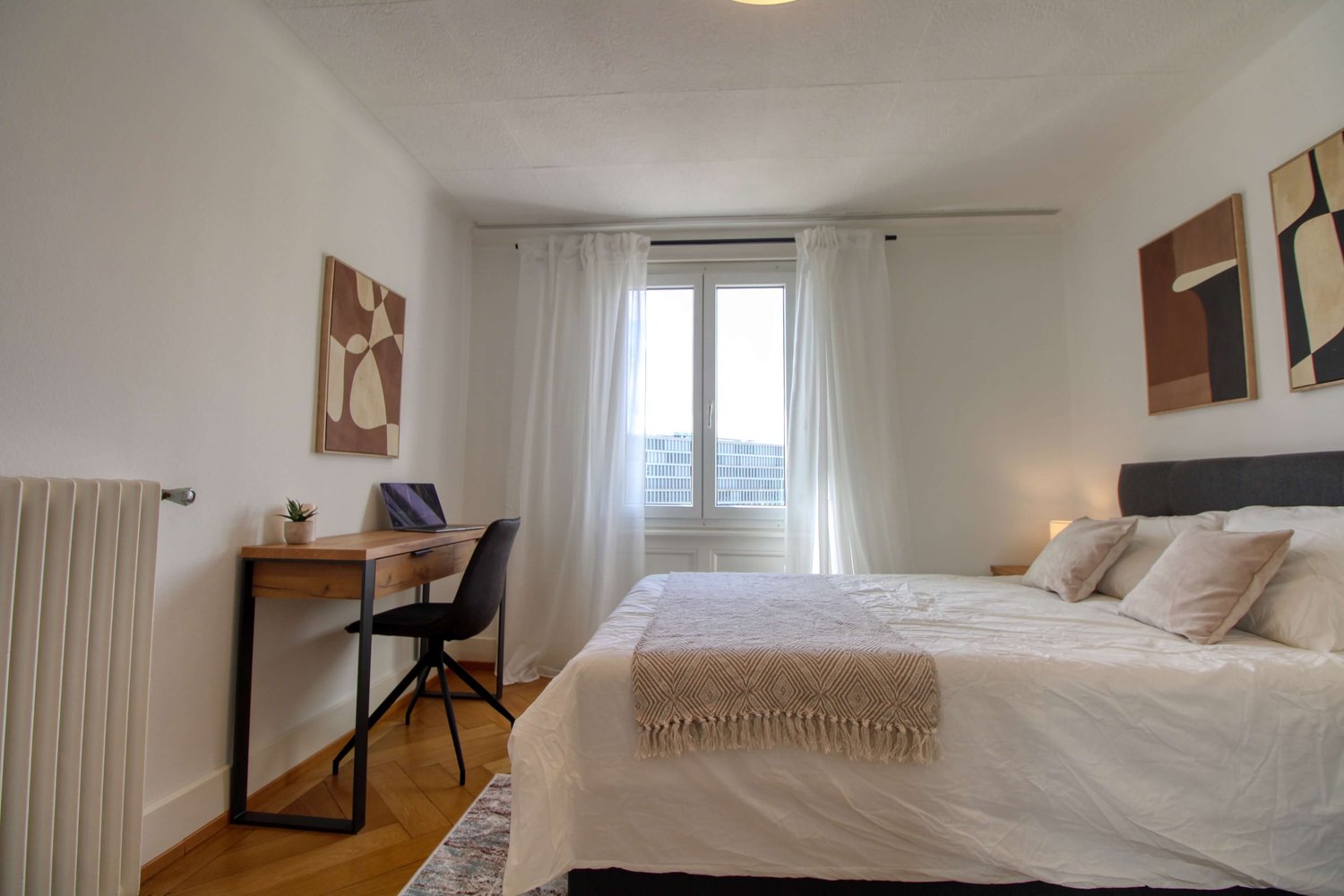 white walls, wooden floors, bed, desk, window with curtains, paintings, lamp, throw blanket