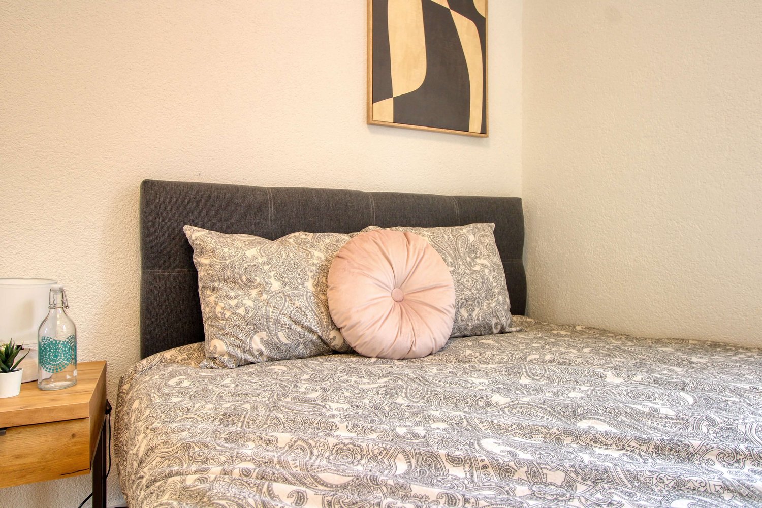 small bed, gray bed headboard, two pillows, paisley patterned sheet, nightstand with plant, picture frame