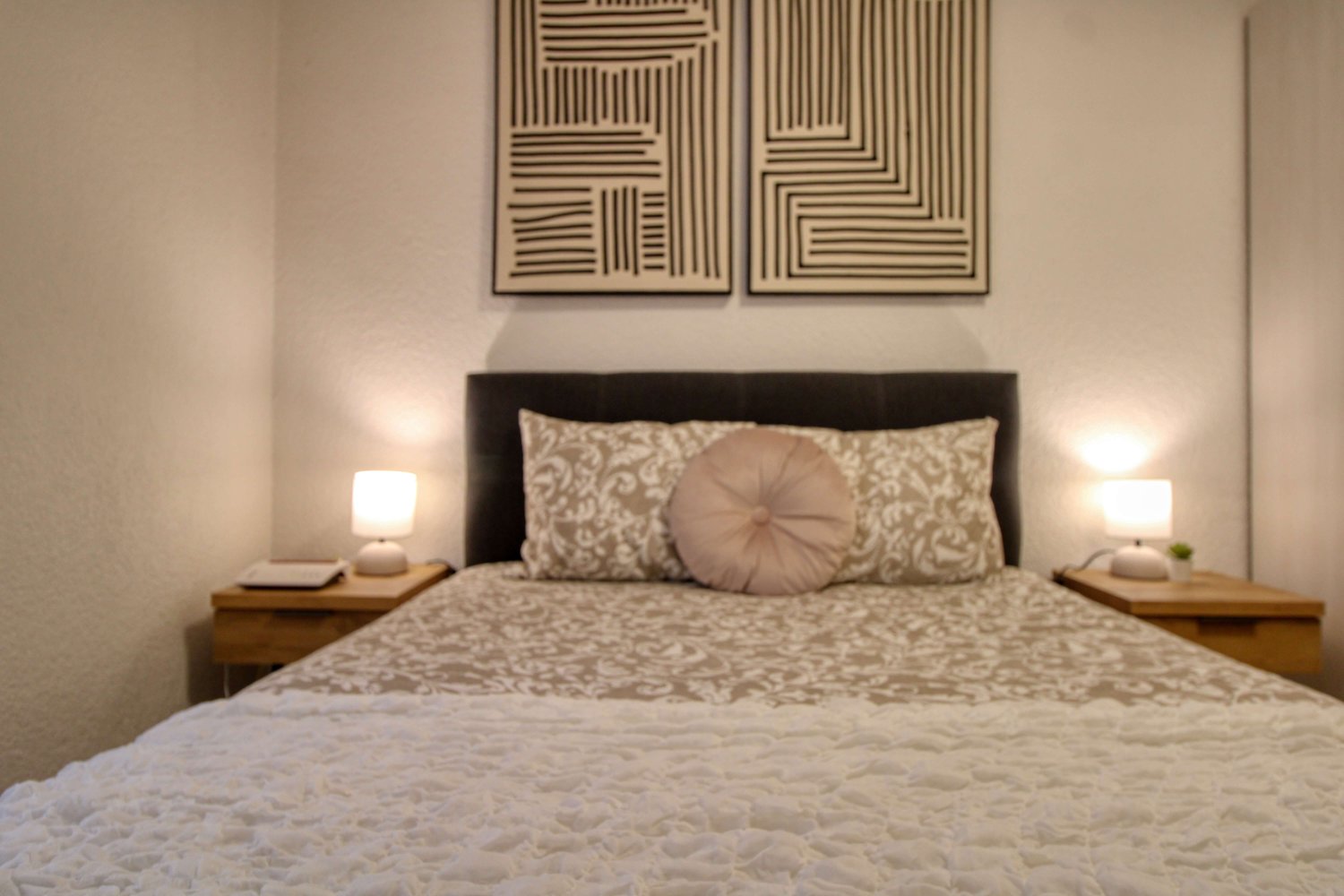 Double bed with headboard, 2 nightstands with lamps, decorative pillow, two decorative wall art pieces, white quilted bedspread