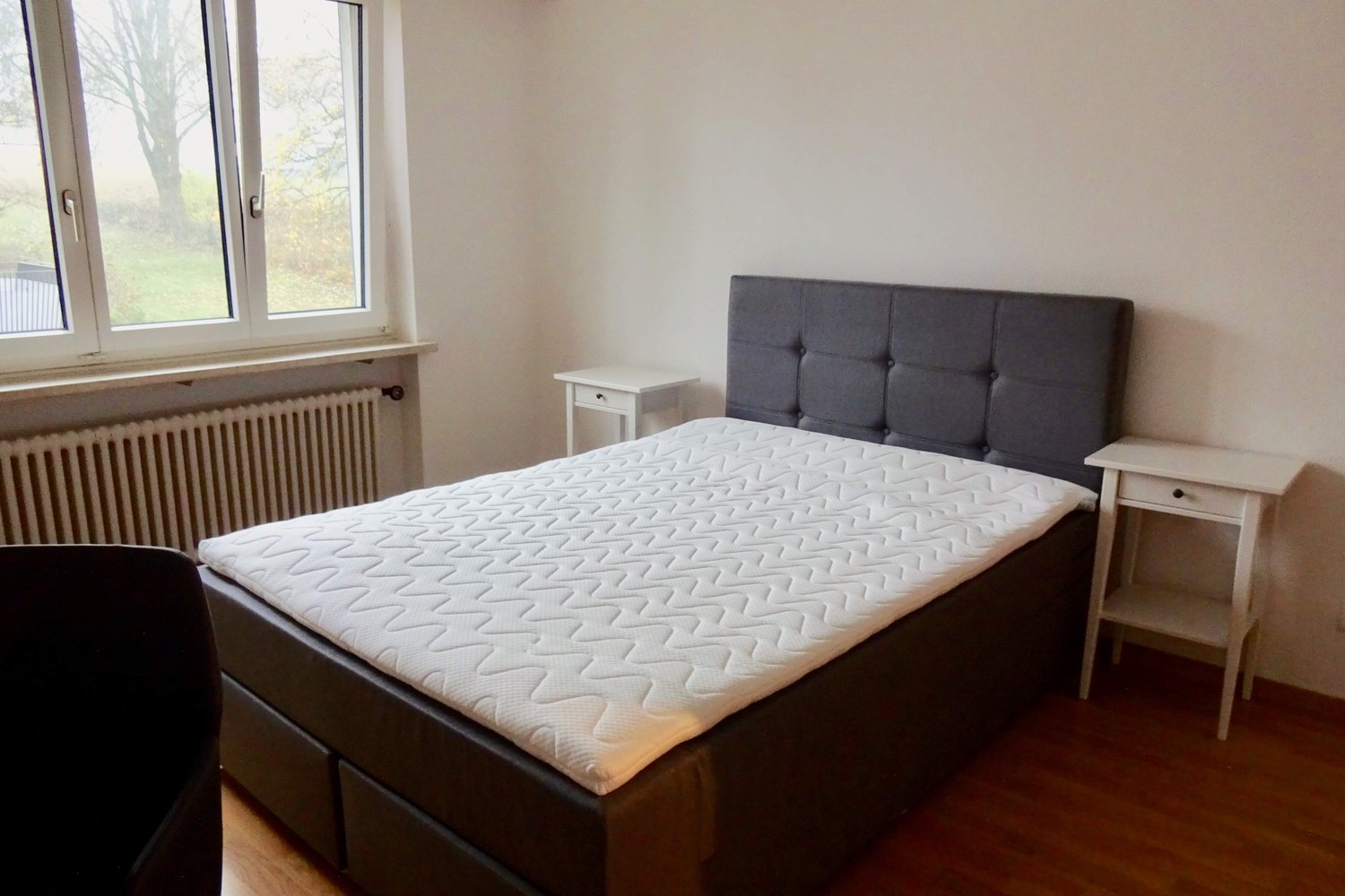 A bed with a white mattress, a bed headboard with a grey fabric cover, white nightstands, a chair, a radiator, and a window with a white frame.