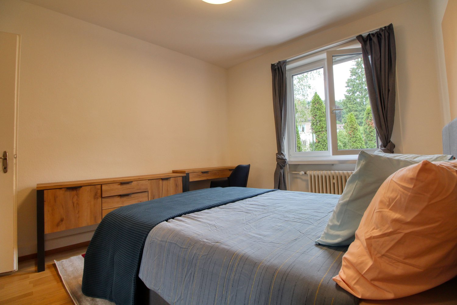 Bed with gray comforter, 2 blue pillows, 1 pink pillow, brown desk, chair, radiator, wooden cabinet, carpeted floor, white wall, glass window with view outside