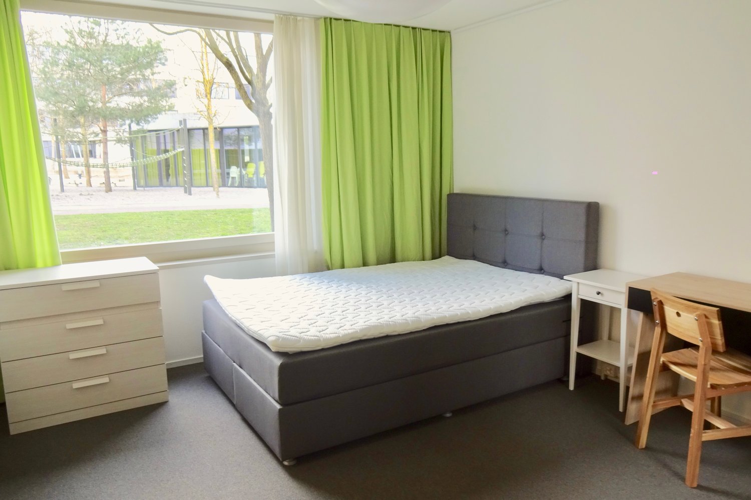 small single bed, bedside table, wooden desk, wooden chair, white dresser, green curtains, window, parquet flooring