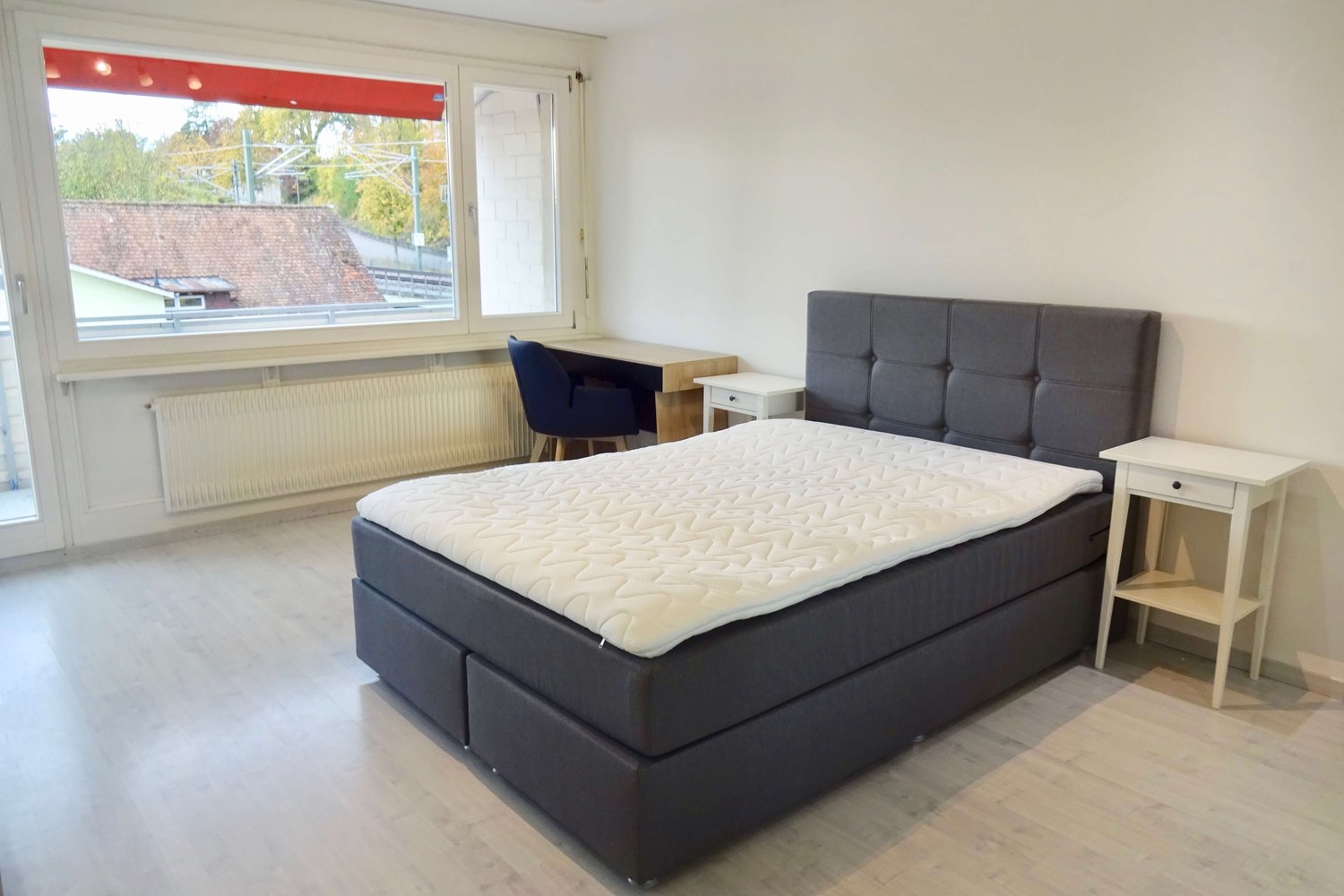 Room with a double bed, desk, and chair by the window, wooden floor, wooden nightstands, white walls