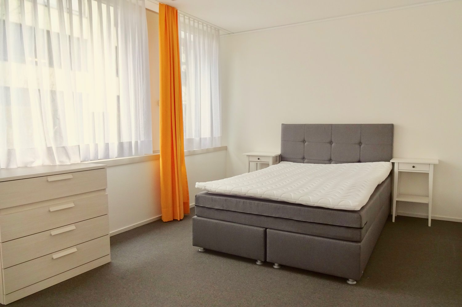 gray bed, gray bed headboard, gray drawers underneath bed, nightstand, white wardrobe, window with white and yellow curtains