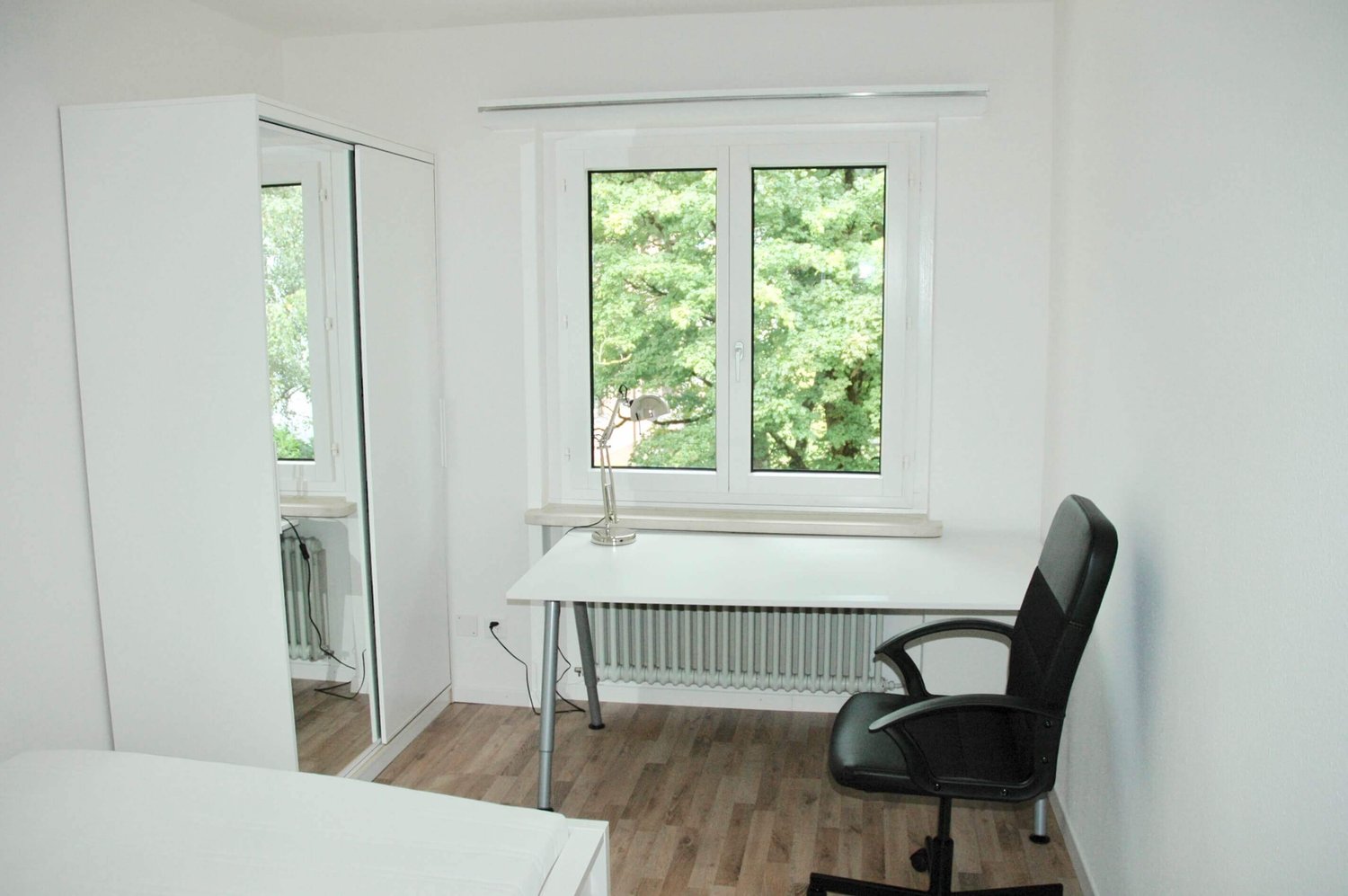 Minimalist bedroom with white walls, wooden floor, bed on the left, desk with chair on the right, large window, closet, desk lamp, and radiator under the window.